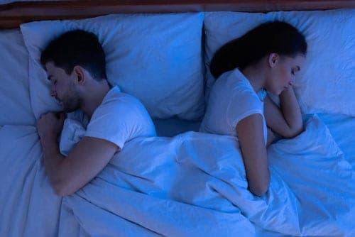 Man and woman in white tops lying back-to-back under white bedding in a dim blue-lit bedroom