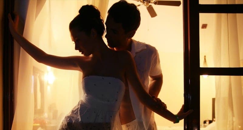 Warm golden-lit silhouette of a woman in a white strapless dress standing in a doorway with a man behind her