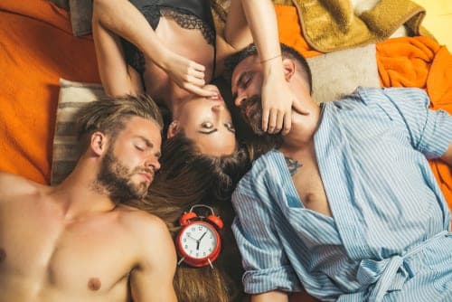 Overhead view of a woman lying between two shirtless men on orange bedding with a red alarm clock placed beside them
