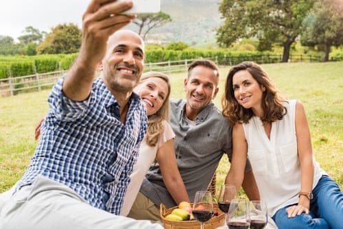 Four smiling adults taking a group selfie at an outdoor vineyard picnic with wine glasses and a fruit basket