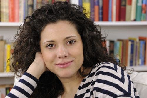 Smiling woman with curly dark hair in a striped sweater resting her chin on her hand in front of a bookshelf