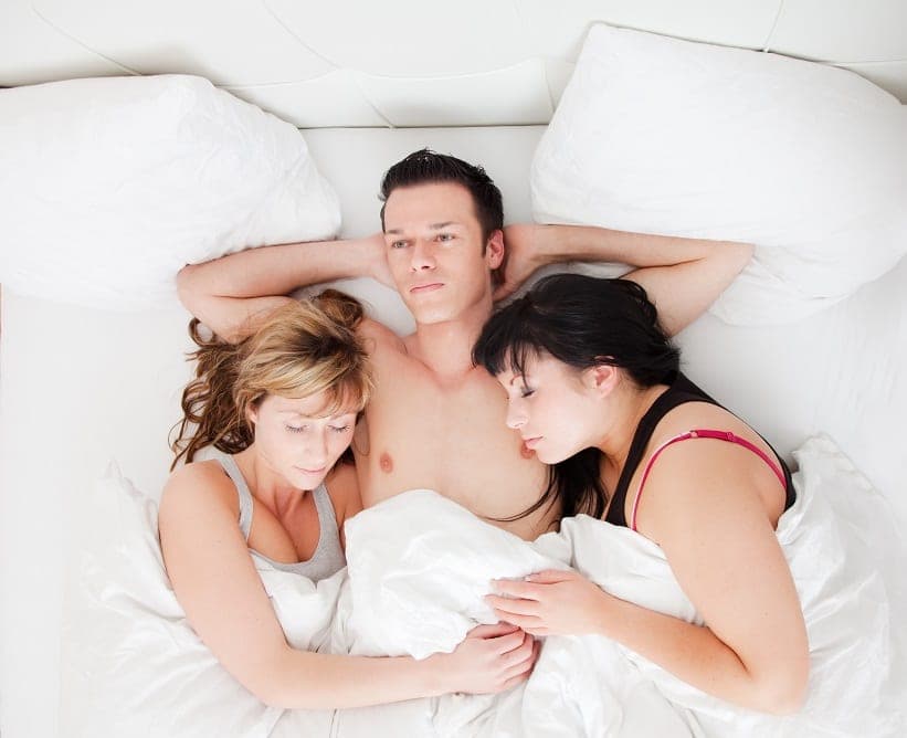 Overhead view of a man between two women on white pillows and sheets, hands behind his head