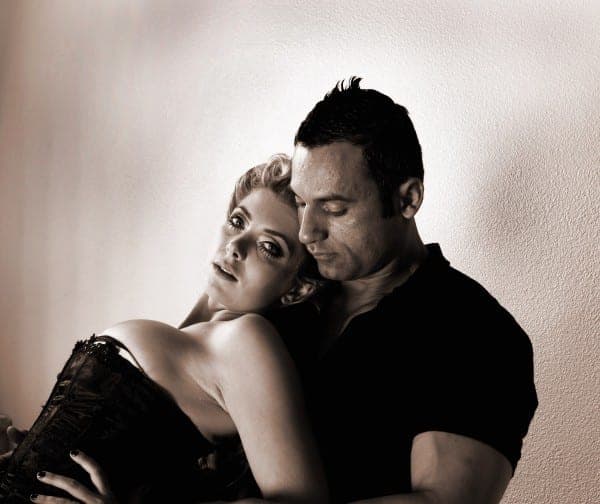 Sepia-toned portrait of a blonde woman in a corset resting her head on a dark-haired man's shoulder