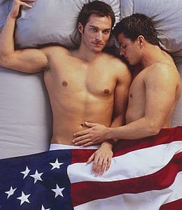 Two shirtless men lying close together on white pillows with an American flag draped across their laps
