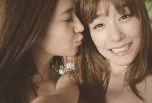 Soft-focus portrait of two young women with long brown hair, one kissing the other's cheek as she smiles