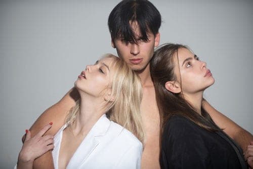 Studio shot of a dark-haired man standing between a blonde woman in white and a brunette in black