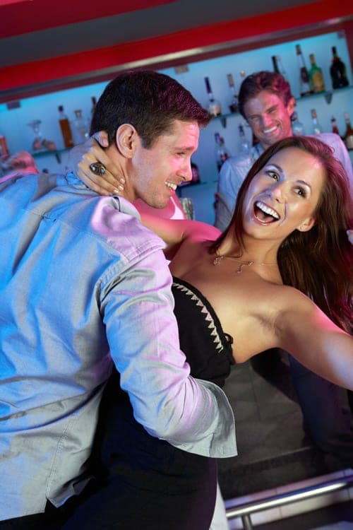 Woman in a black dress laughing as she dances with a man in a grey shirt at a neon-lit bar