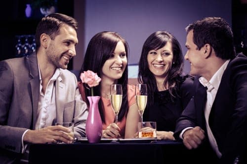 Two couples sharing drinks and smiling around a small table with a pink rose and champagne flutes