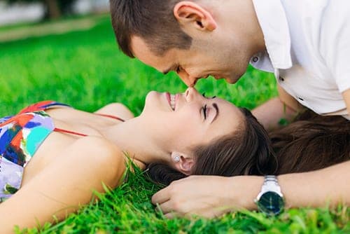 Young couple lying on green grass nose to nose, smiling at each other in bright sunlight