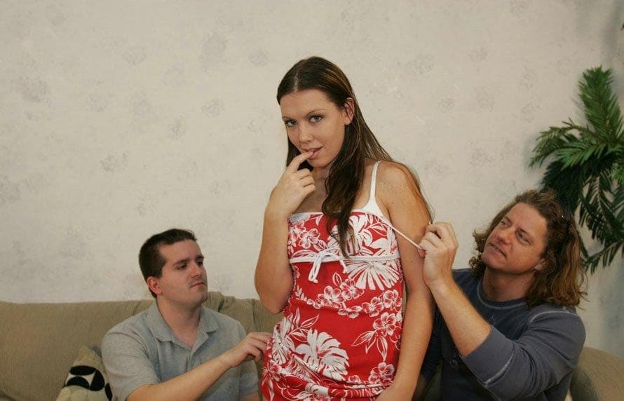 Brunette in a red floral sundress stands between two seated men who tug at her dress straps