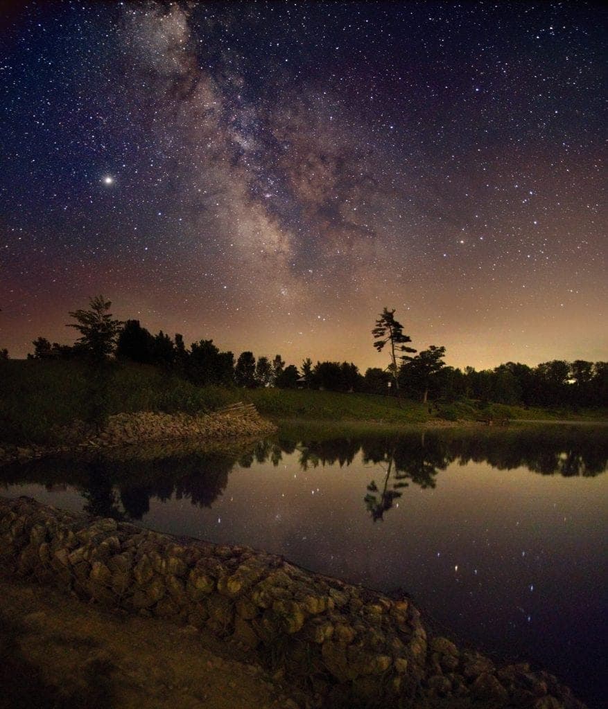 Milky Way galaxy arching over a calm lake reflecting stars, with silhouetted trees on the shoreline