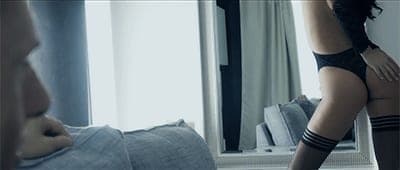 Dim bedroom scene with a man lying on a bed watching a woman in black lingerie and thigh-high stockings