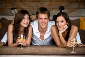 Two women and a man lying on a couch smiling at the camera, the women holding glasses of white wine