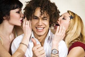 Curly-haired man giving a thumbs up while two women lean in to kiss his cheeks