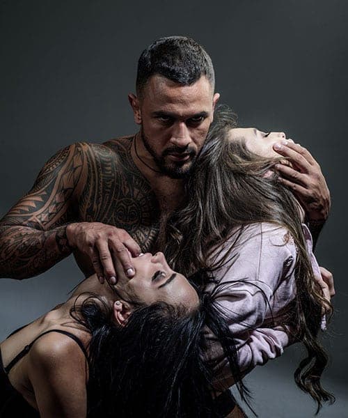 Studio portrait of a tattooed shirtless man holding two women with long dark hair in a dramatic pose