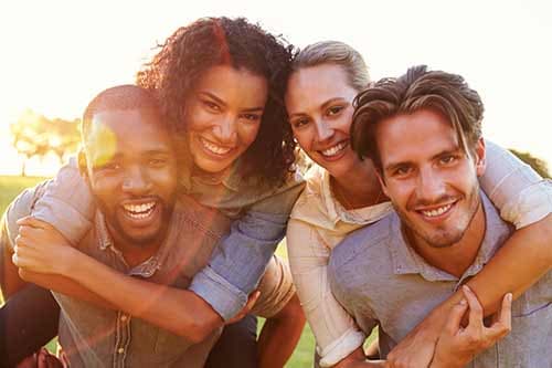 Two couples smiling at the camera outdoors at sunset, the women giving piggyback rides to the men