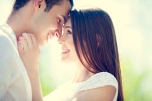 Young couple in white shirts touching foreheads and smiling closely against a sunlit outdoor backdrop