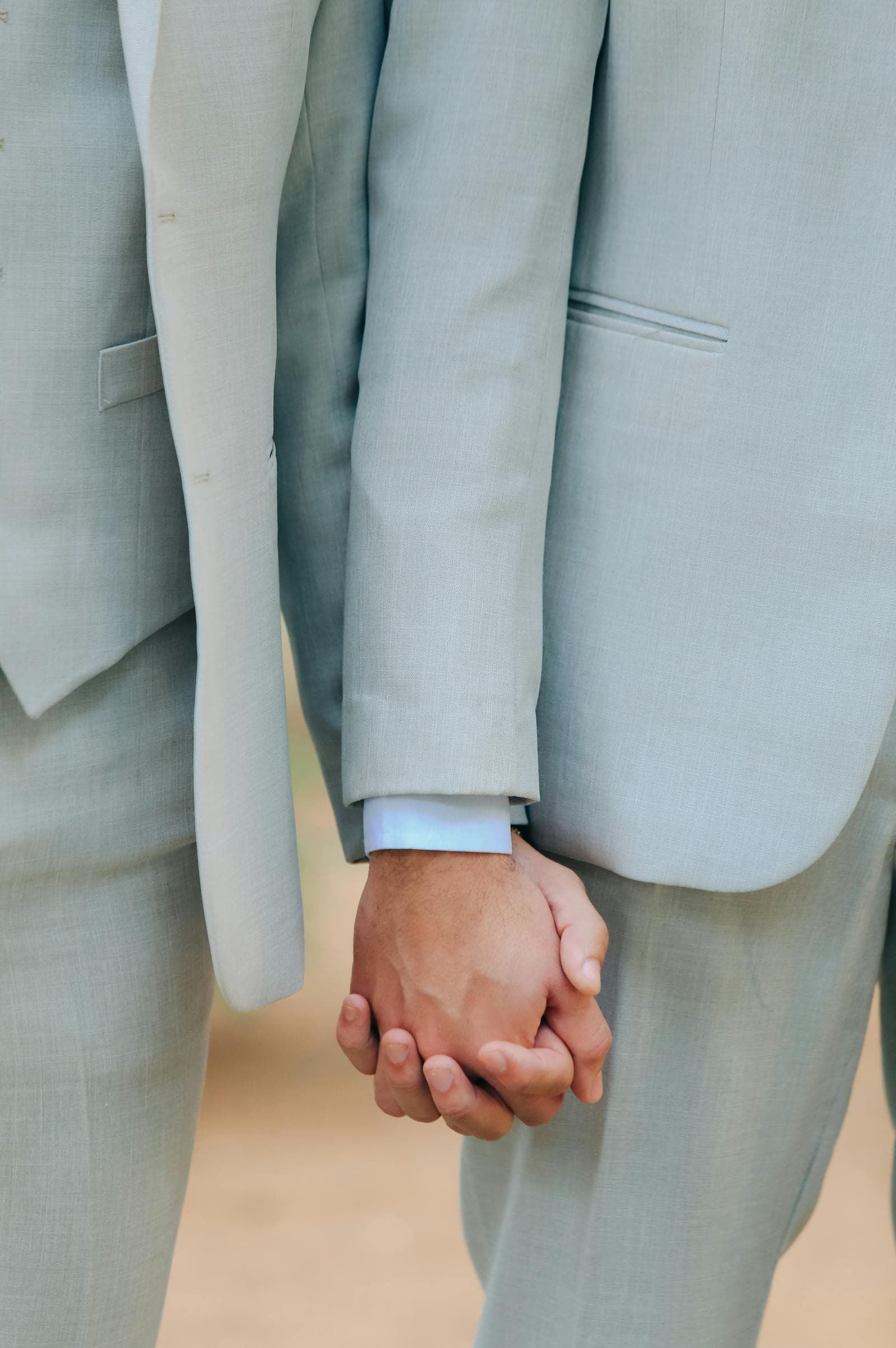 Close-up of two men's hands intertwined, both wearing pale blue suits, in a quiet gesture of connection