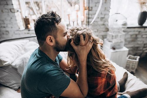 Bearded man in teal shirt kissing a laughing woman's forehead on a bed in a cozy string-lit bedroom