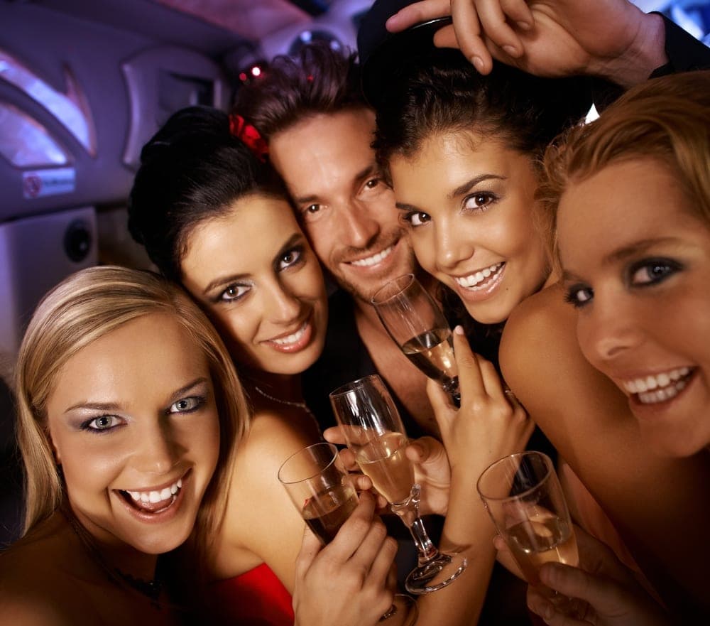 Group of smiling friends in a limousine leaning together and raising champagne flutes at the camera