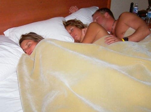 Three people sleeping under a yellow blanket in a hotel bed, two women and one man resting close