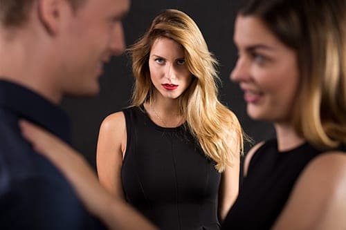 Blonde woman in black dress watches with a tense expression as a man and brunette woman flirt nearby
