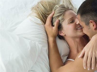 Couple lying in white bedding, woman smiling with eyes closed as a dark-haired man leans in close