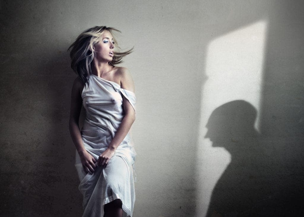 Blonde woman in a loose white dress leans against a concrete wall as a shadowy male silhouette looms beside her