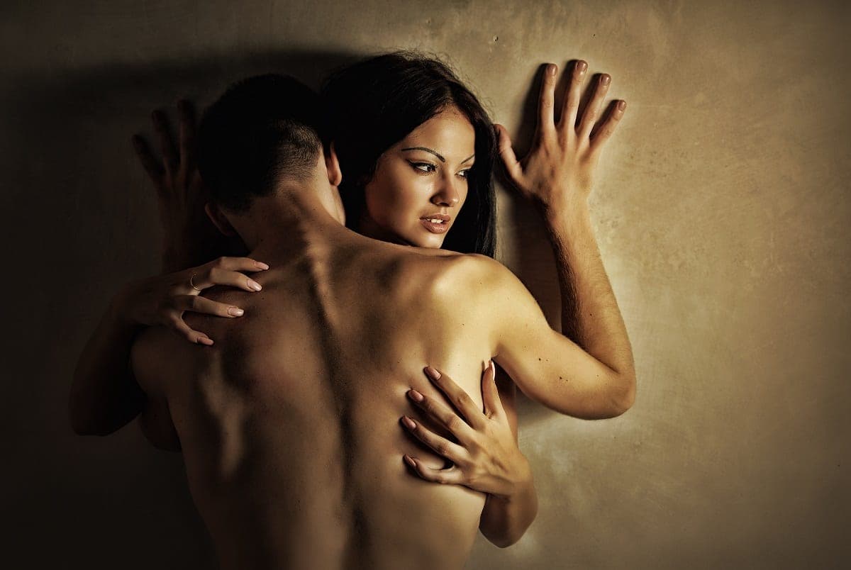Brunette woman pressed against a warm-lit wall embracing a shirtless man, hands splayed against the surface