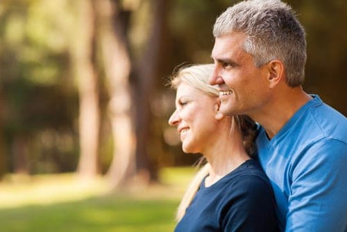 Grey-haired couple in blue shirts smiling together outdoors with blurred sunlit trees behind them