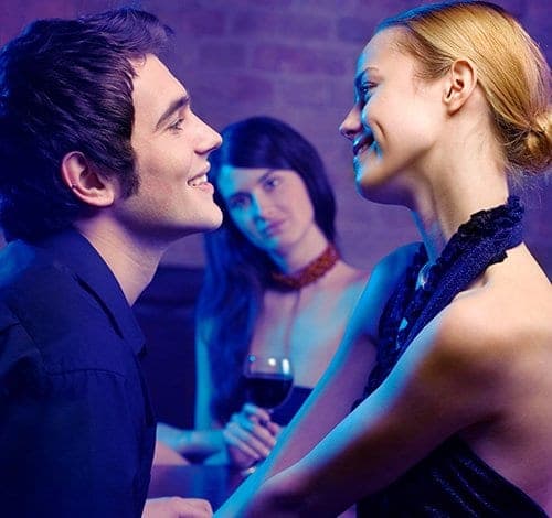 Smiling couple flirting at a bar while a second woman watches them with a wine glass in hand