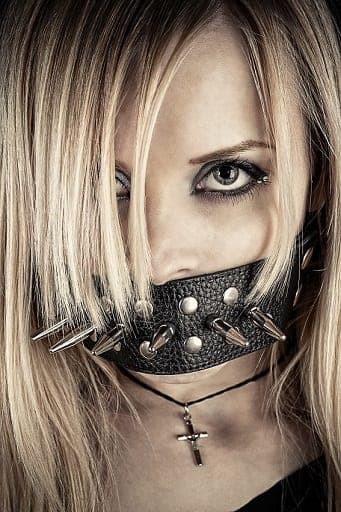 Blonde woman wearing a studded leather gag strap and cross necklace staring at the camera