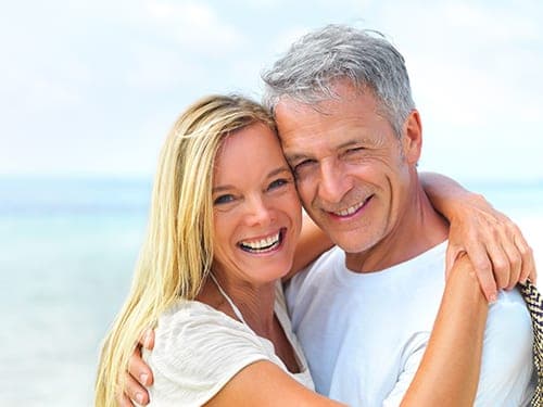 Smiling grey-haired man and blonde woman embracing on a bright beach with ocean behind them