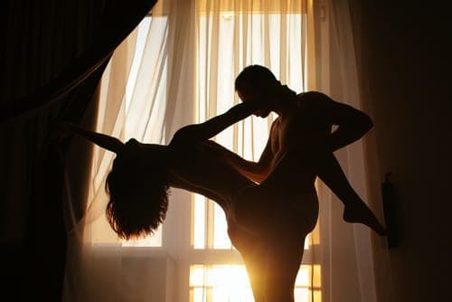 Silhouetted couple embracing in front of sunlit sheer curtains in a softly lit bedroom