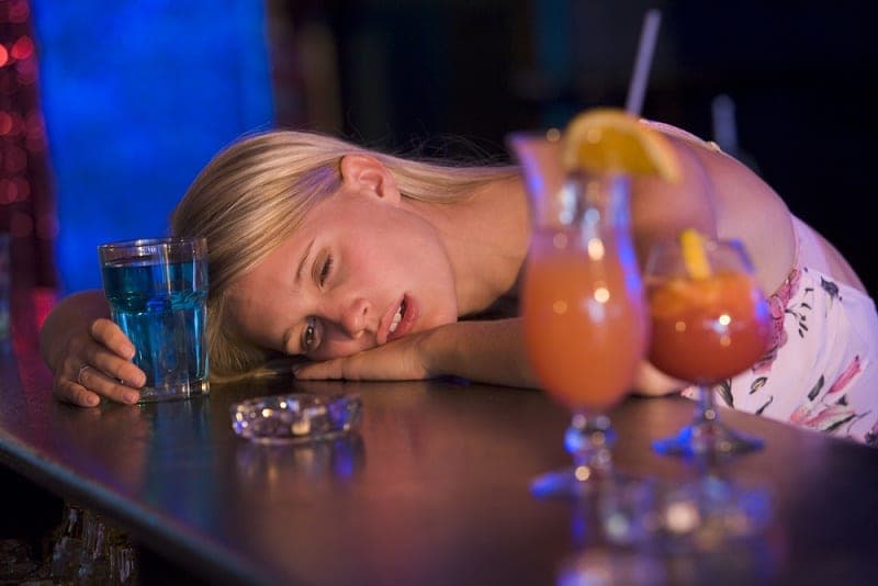 Blonde woman resting her head on a bar counter next to cocktails and an ashtray in blue light