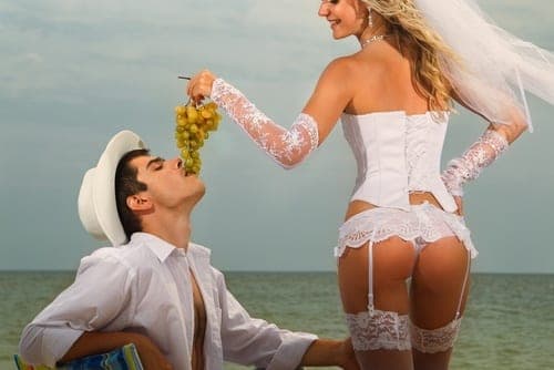 Bride in white corset and veil feeding grapes to a man in a white hat and shirt on a beach