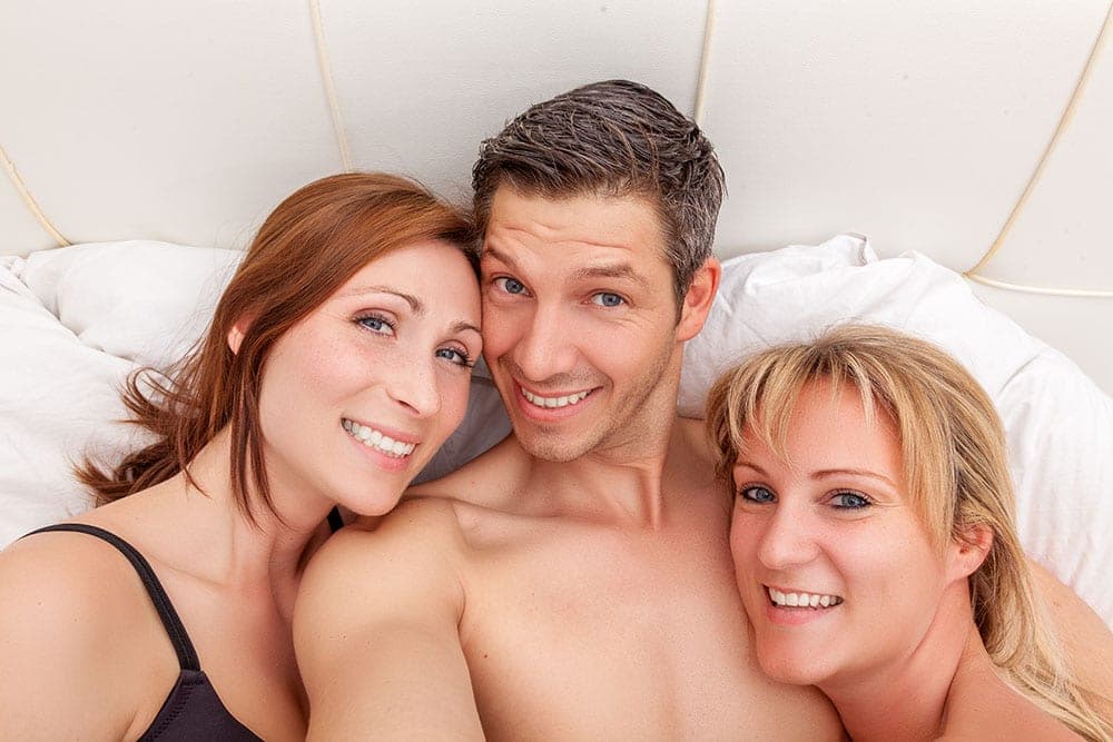 Smiling man between two women lying on white bed pillows, all looking up at the camera