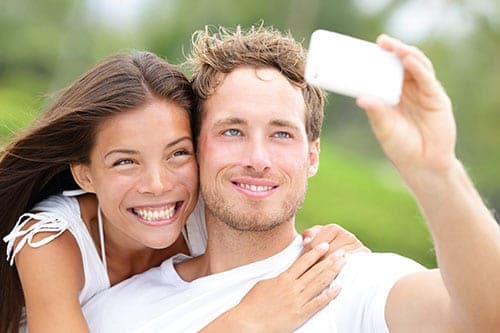 Smiling young couple take a selfie together outdoors, the man holding a white smartphone up close
