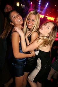 Three young women in short dresses laughing and hugging on a crowded nightclub dance floor