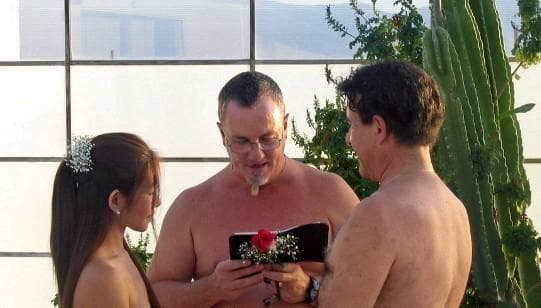 Outdoor nude wedding ceremony with officiant reading from a book between a bride and groom holding a rose