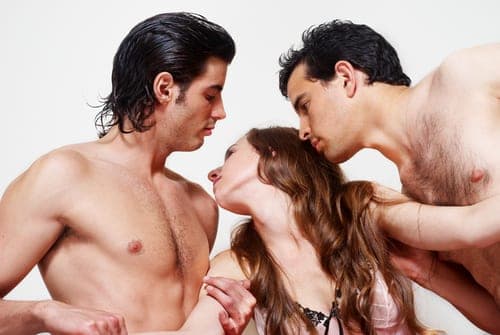 Two shirtless men lean in close to kiss a woman between them against a plain white background