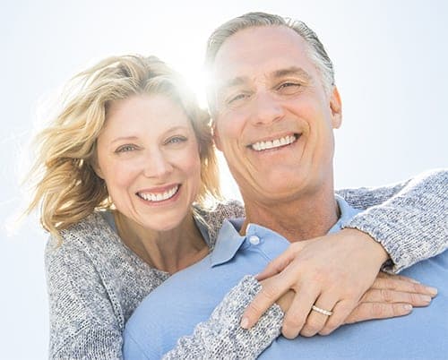 Smiling middle-aged couple embrace outdoors in bright sunlight, both wearing grey and blue sweaters