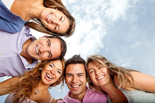 Five young adults huddled in a close circle viewed from below, smiling down at the camera under a blue sky