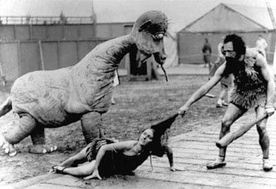 Vintage black-and-white film still of costumed cavemen and a dinosaur prop dragging a woman on a set