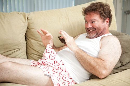 Bearded man in a white tank top and heart-print boxers grins while texting on a couch