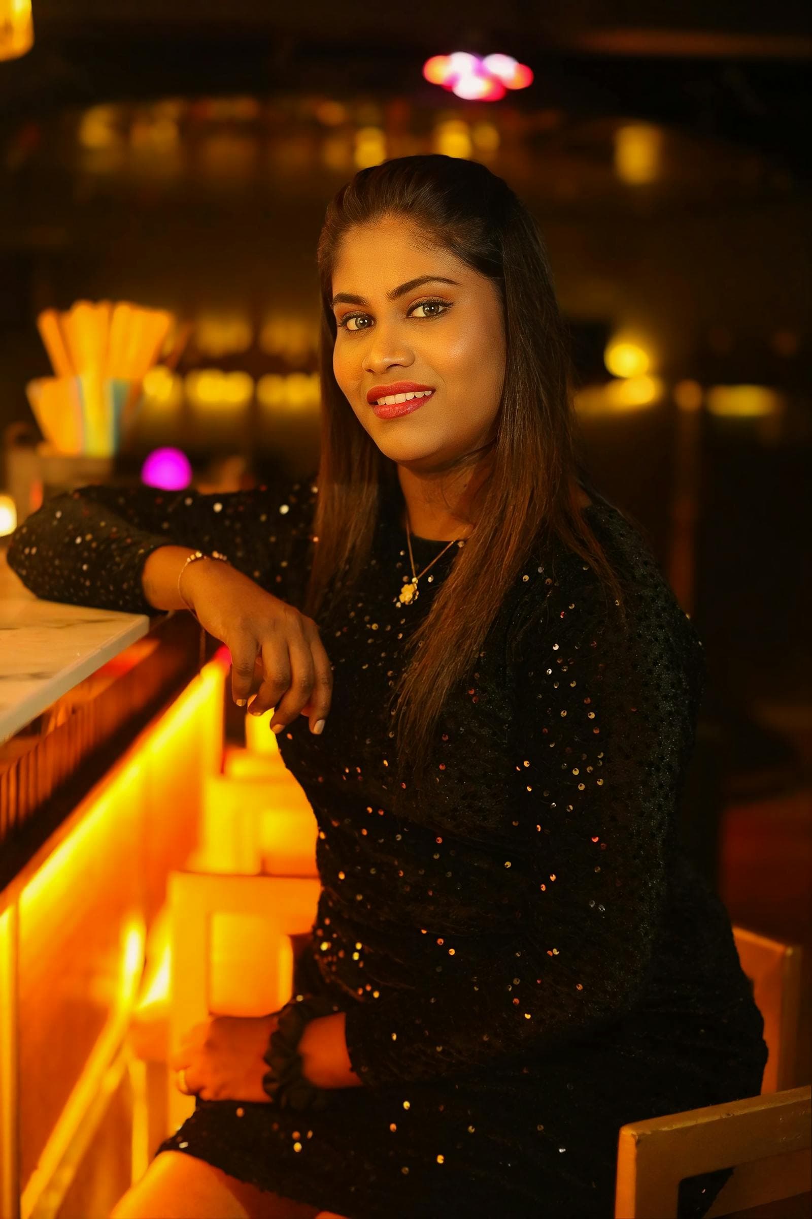 Woman in a black sequined dress at a marble bar counter, warm amber lights and softly blurred bottles glowing behind her