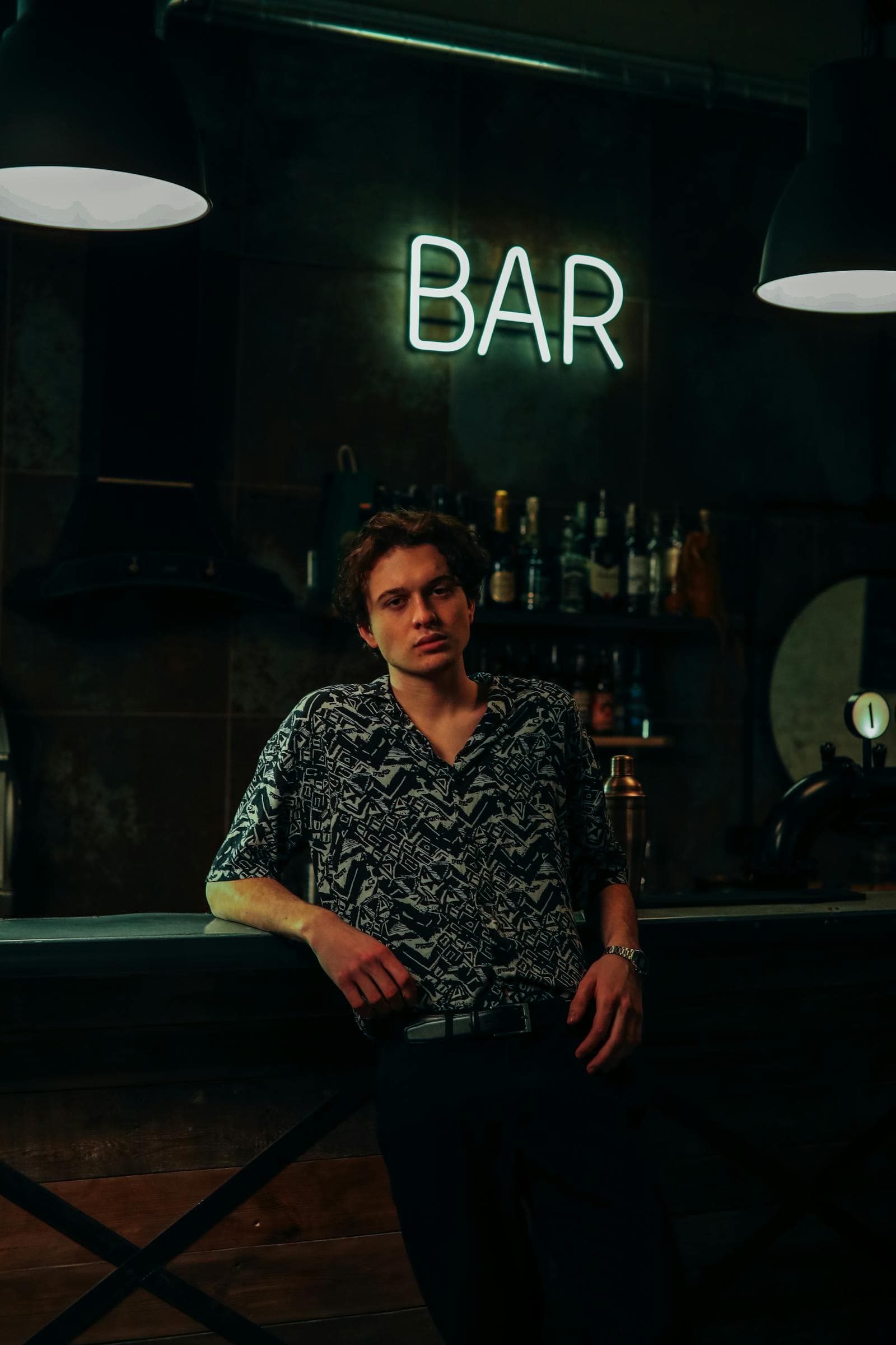 Man in a dark patterned shirt leaning against a bar counter beneath pendant lights and a glowing neon BAR sign