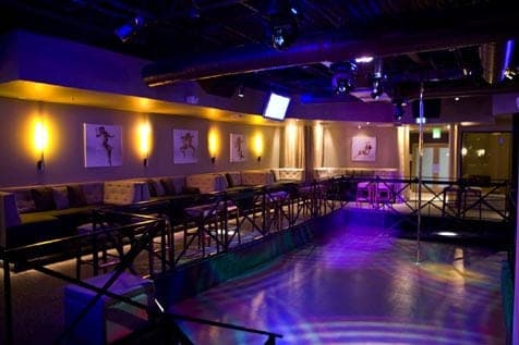 Empty nightclub interior with a dance floor lit by purple lights, stripper pole, and lounge seating
