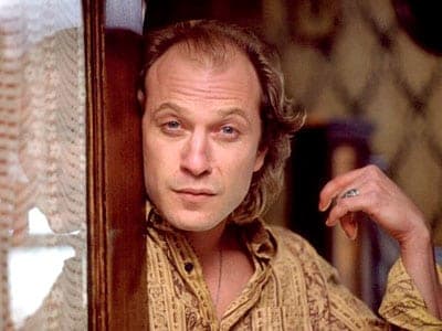 Man with curly reddish hair in a patterned shirt leans against a doorframe holding a small object