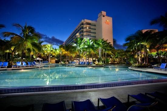 Nighttime resort pool surrounded by palm trees and lounge chairs with a lit hotel tower behind